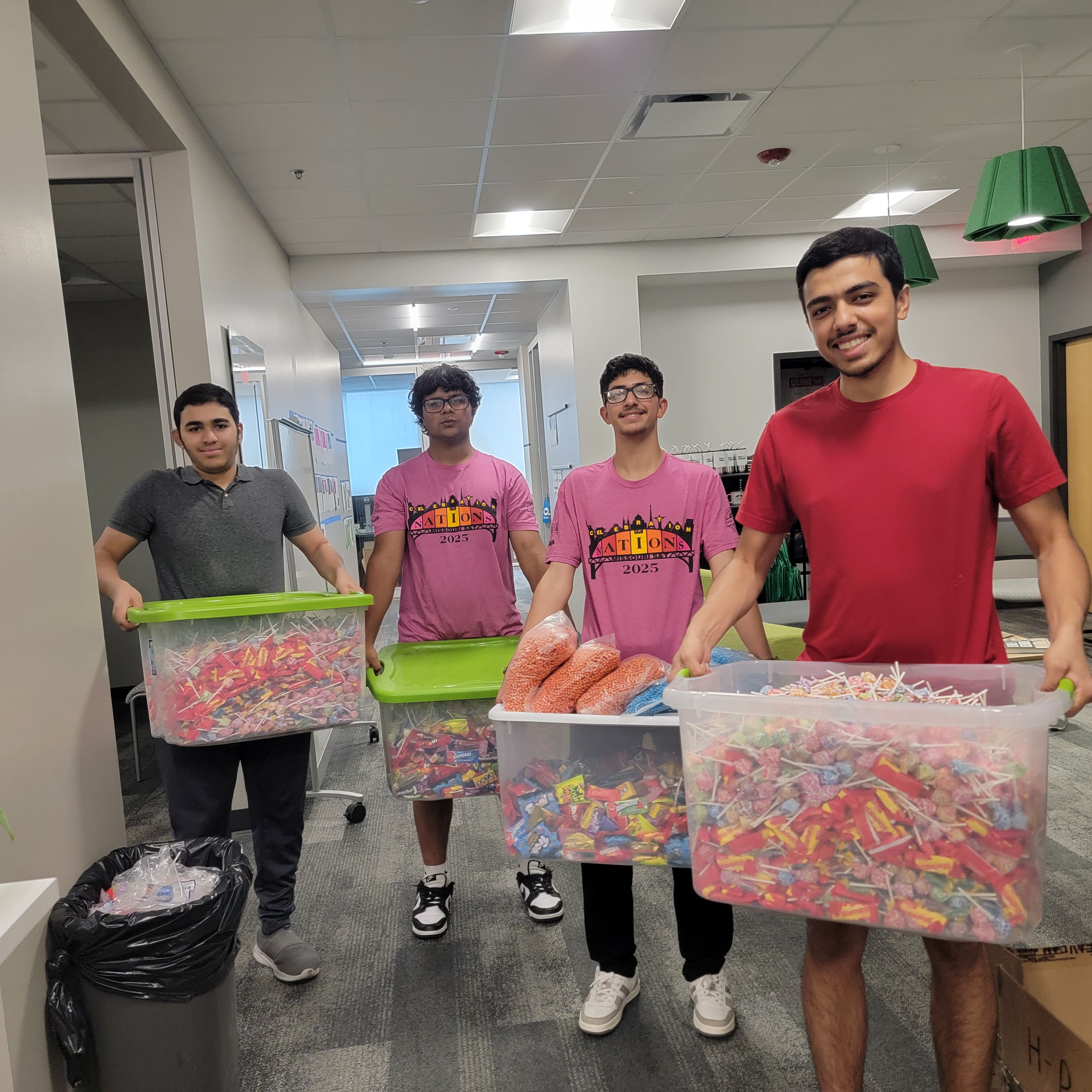 Four young men stand in a hallway, each holding a large bin filled with bags of