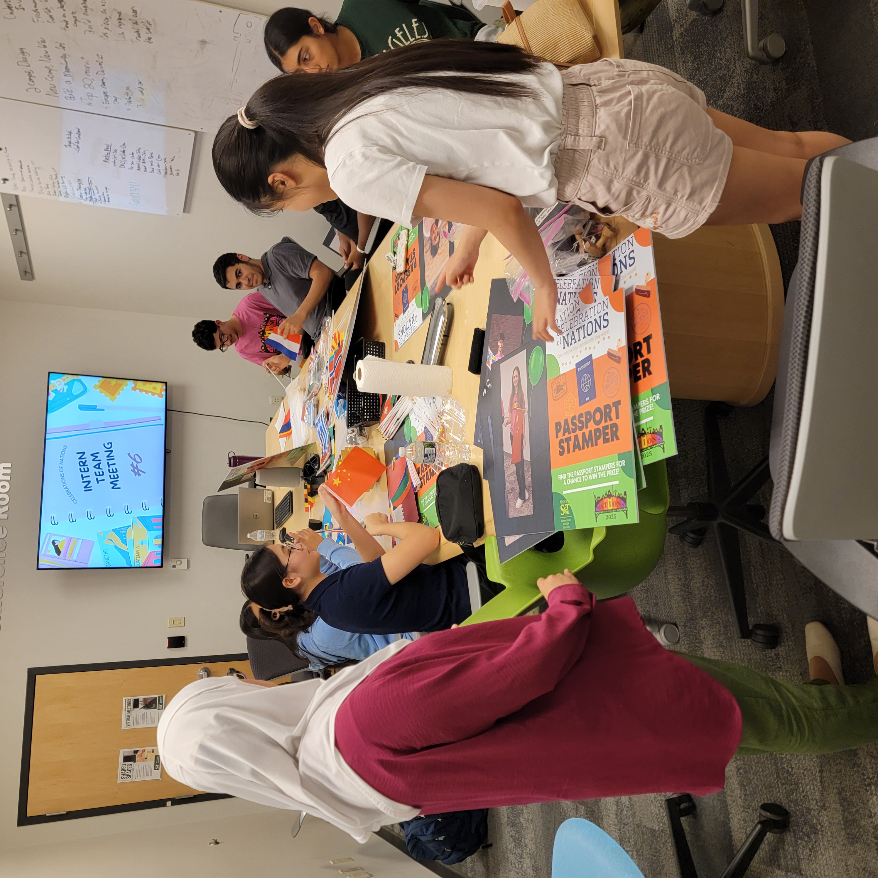 A diverse group of interns gathers around a table covered in colorful posters and stationery during a team meeting. A screen displays 