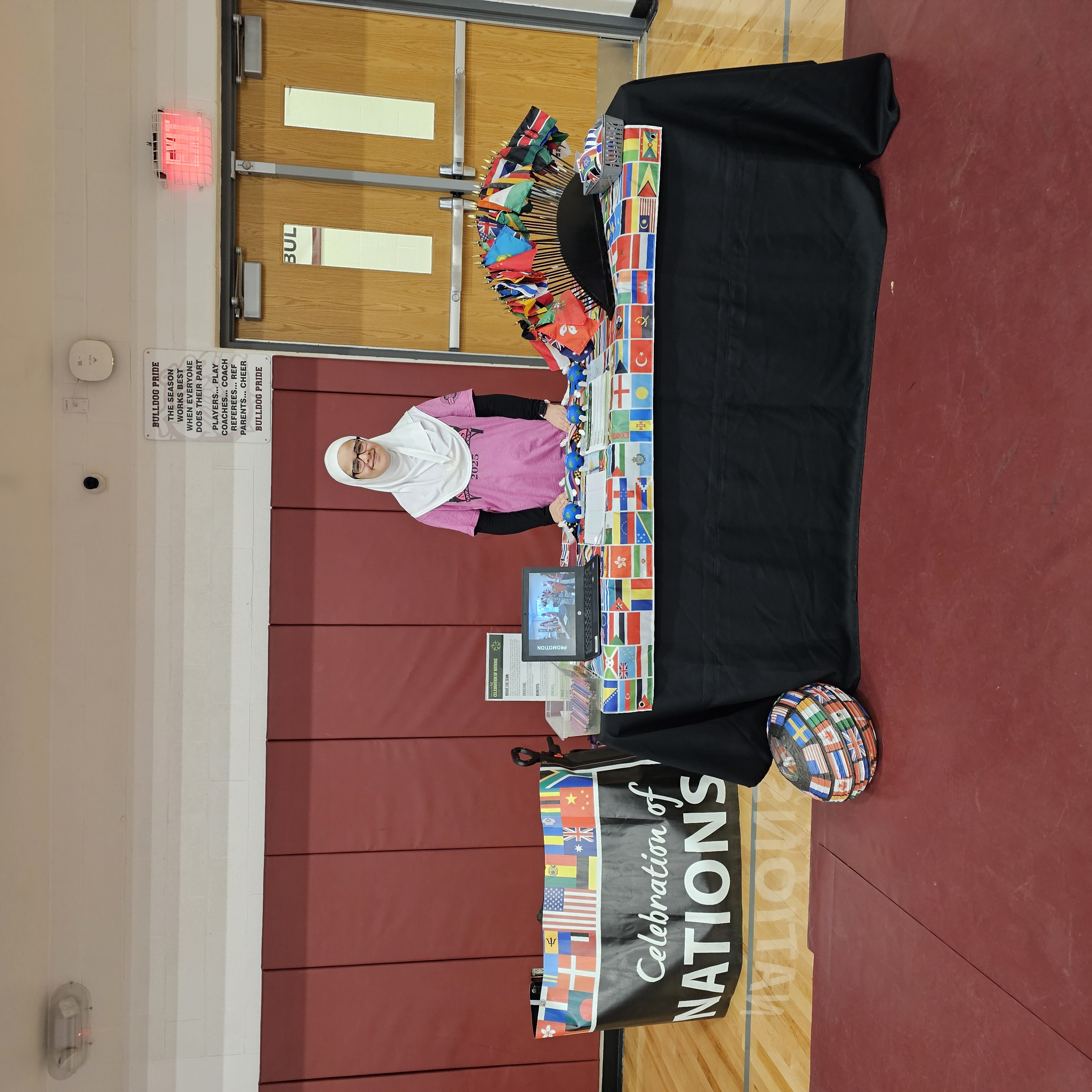 A girl stands smiling behind a table covered with various international flags in a school gym. A 