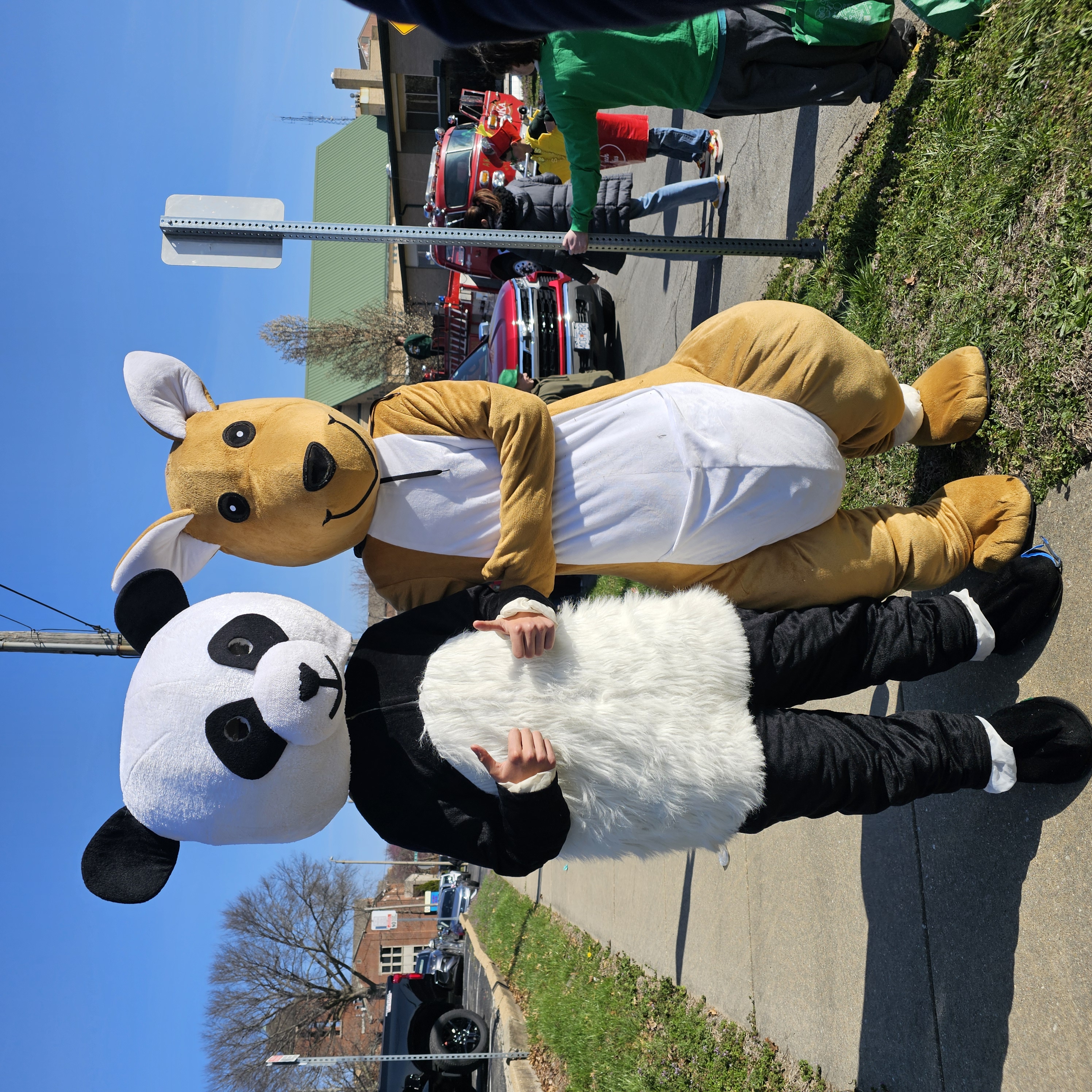 Two people in panda and kangaroo costumes pose cheerfully on a sunny sidewalk
